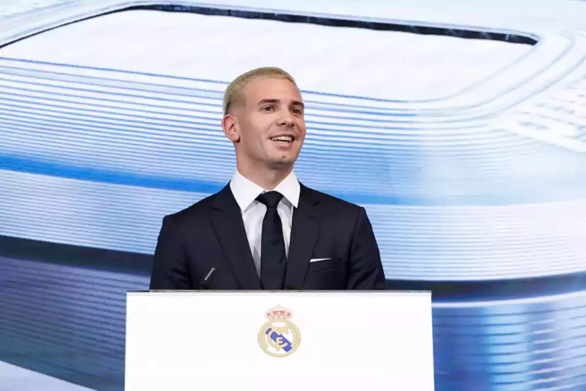 Un hombre de cabello corto y rubio vestido con traje oscuro y corbata negra sonríe mientras habla en un podio con el escudo del Real Madrid al frente y un fondo azul claro.