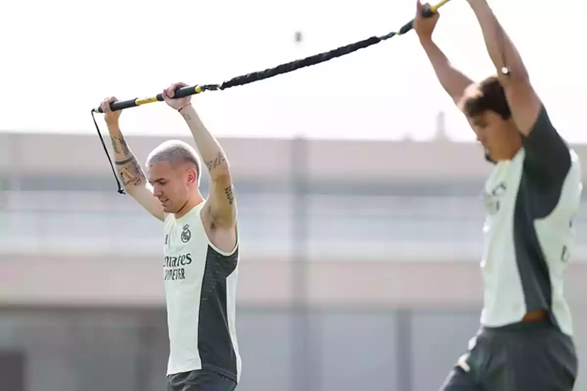 Dos futbolistas entrenan al aire libre levantando una barra elástica sobre sus cabezas mientras usan ropa deportiva del Real Madrid