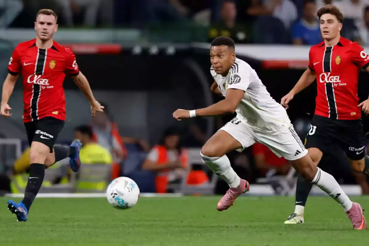 Tres futbolistas disputan el balón durante un partido, dos con uniforme rojo y negro y uno con uniforme blanco, en un estadio lleno de espectadores.