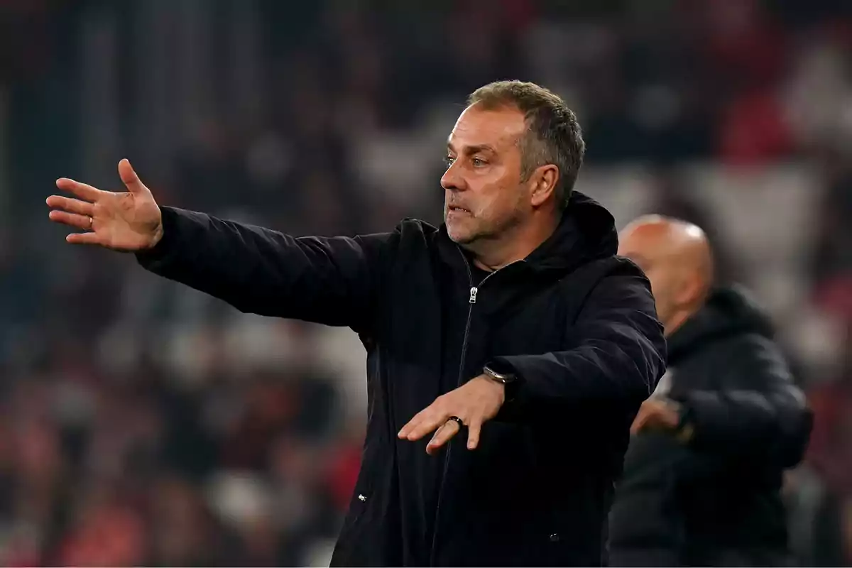 Un hombre con chaqueta oscura gesticula con la mano extendida en un entorno deportivo.