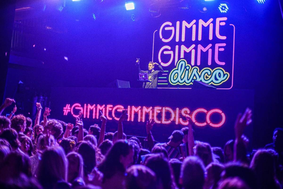 Ambiente vibrante en la fiesta Gimme Gimme Madrid con DJ en cabina y público bailando frente al cartel luminoso.