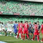 Jugadores del Real Betis y el Sevilla FC saludándose antes del inicio de un Gran Derbi en el estadio Benito Villamarín, con la grada verdiblanca completamente llena al fondo.